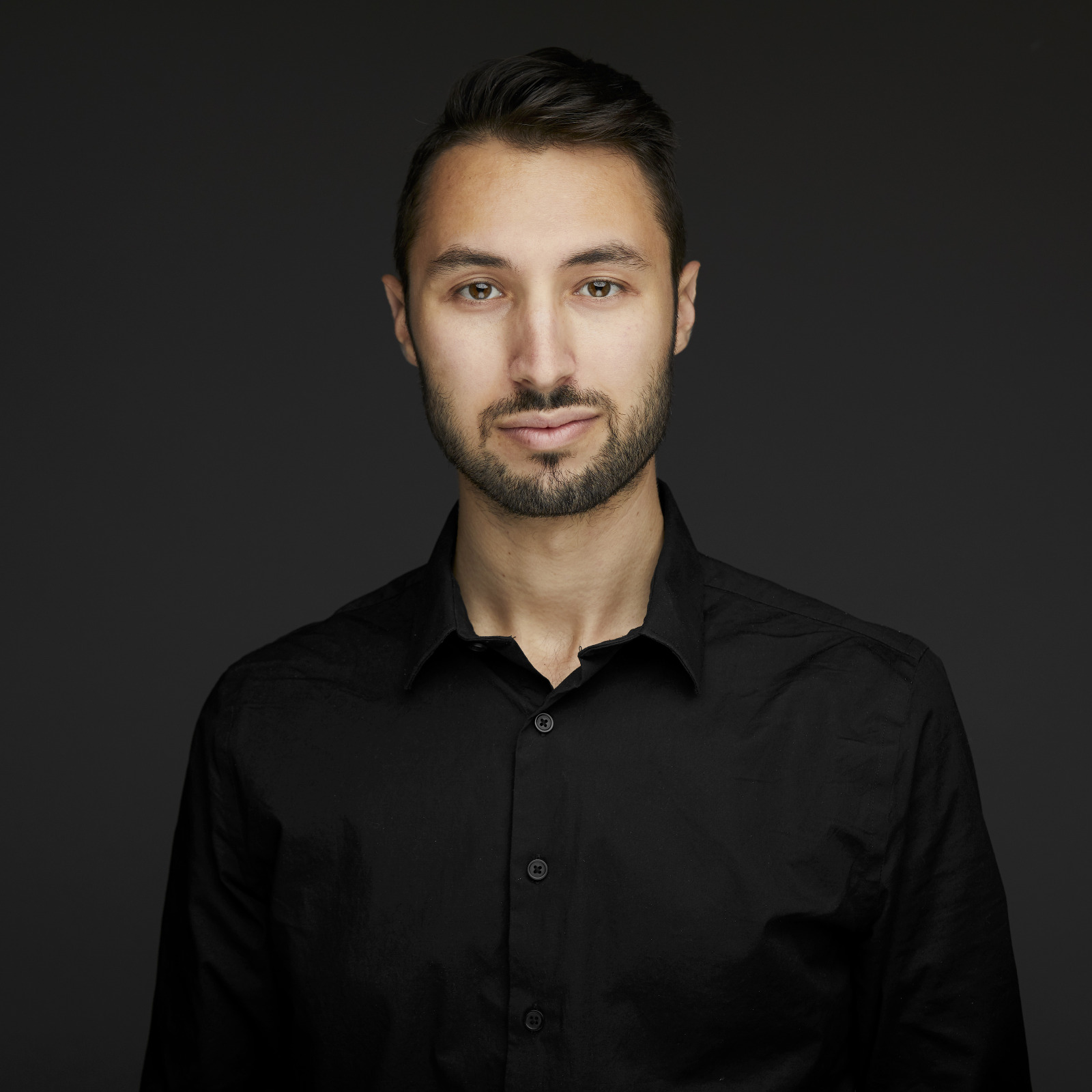 A professional headshot of a young man wearing a black button down