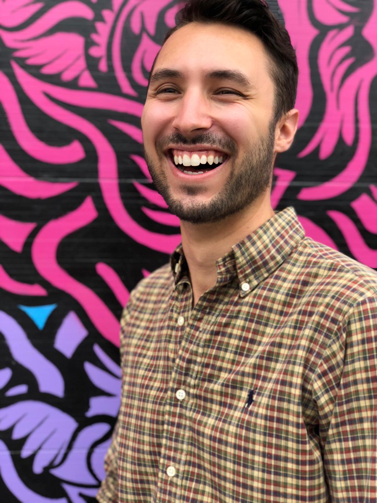 A professional headshot of a young man wearing a black button down